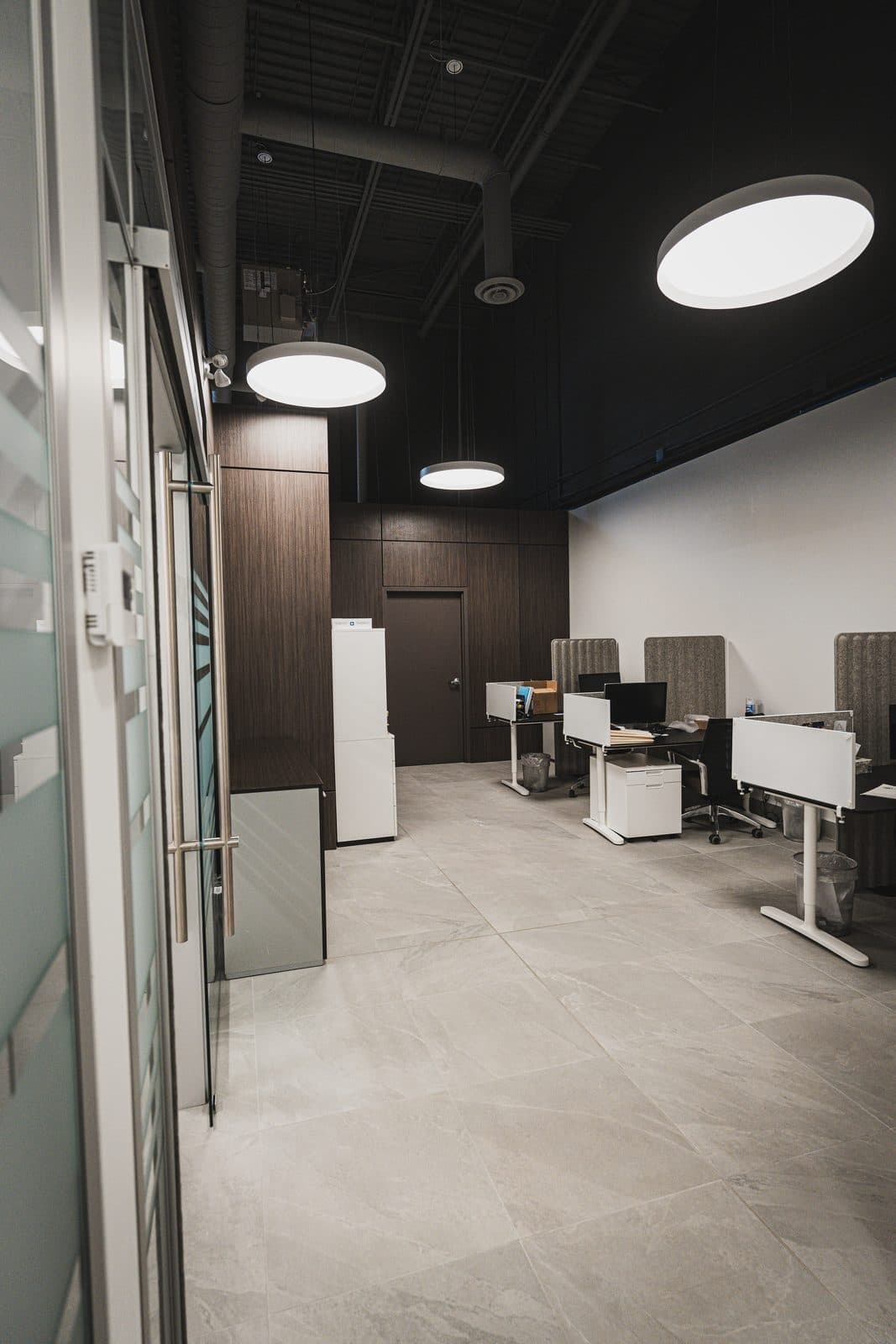 Glazed corridor with striped privacy film opening onto workstations and pendant lighting beneath a black ceiling.