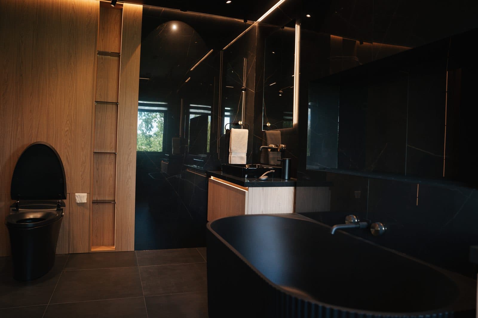 Fluted black freestanding tub between warm oak vanities against dark marble