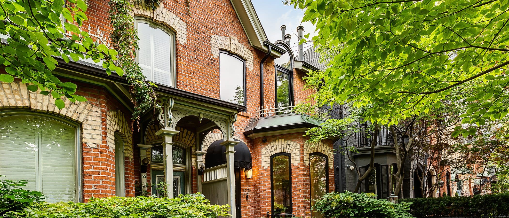 Two story red brick Victorian facade with cream brick banding, arched black framed windows, iron fence, and mature trees along the front walk.