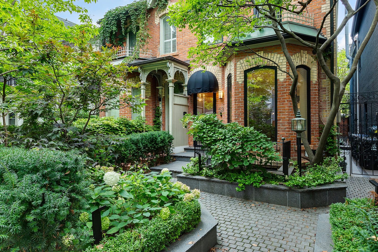 Stone paver walkway leading to a black glass front door beneath a black canopy awning, flanked by hydrangeas and clipped hedges.