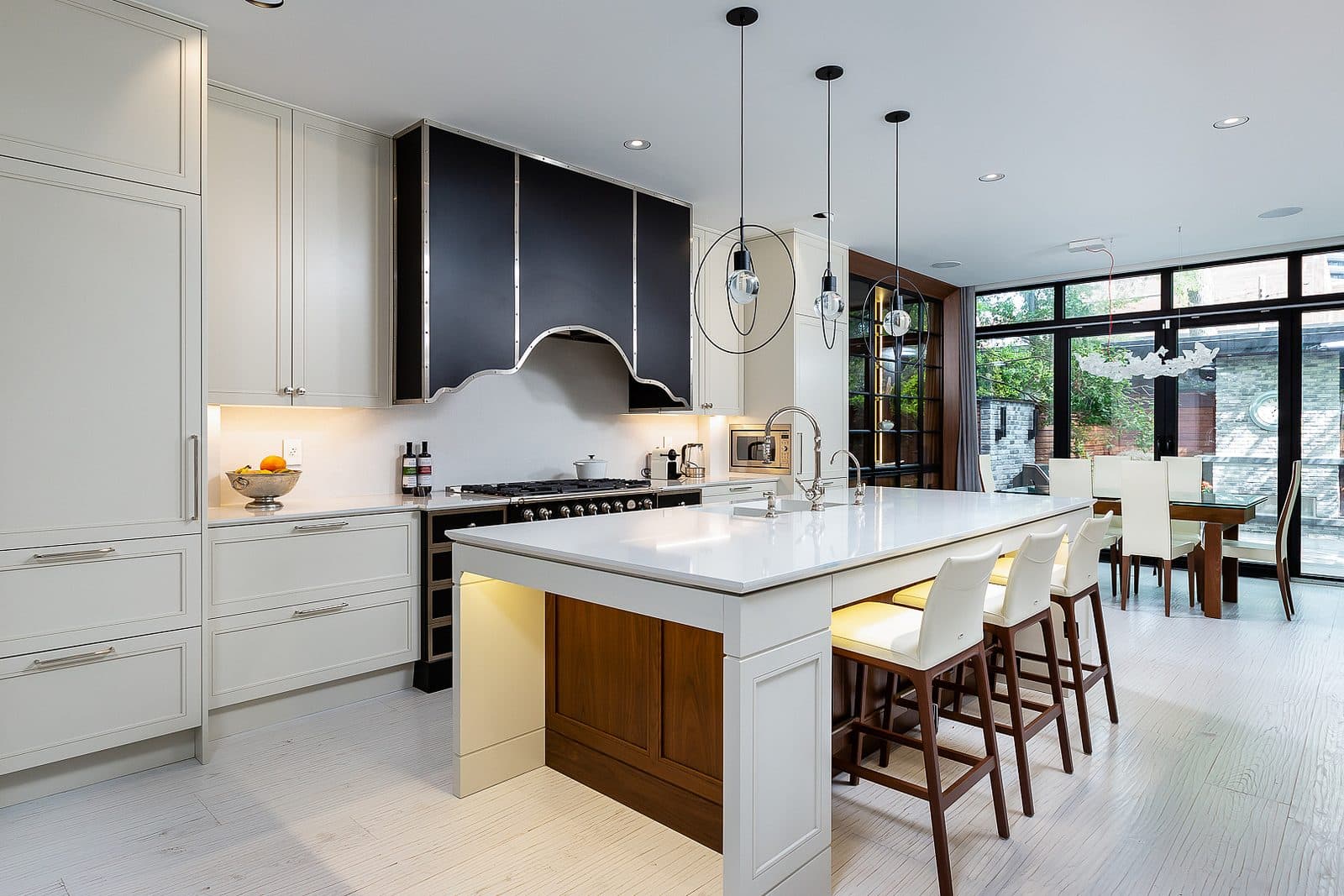 Open concept kitchen with white cabinetry, black paneled range hood, waterfall island, and dining table beyond glass doors to the garden.