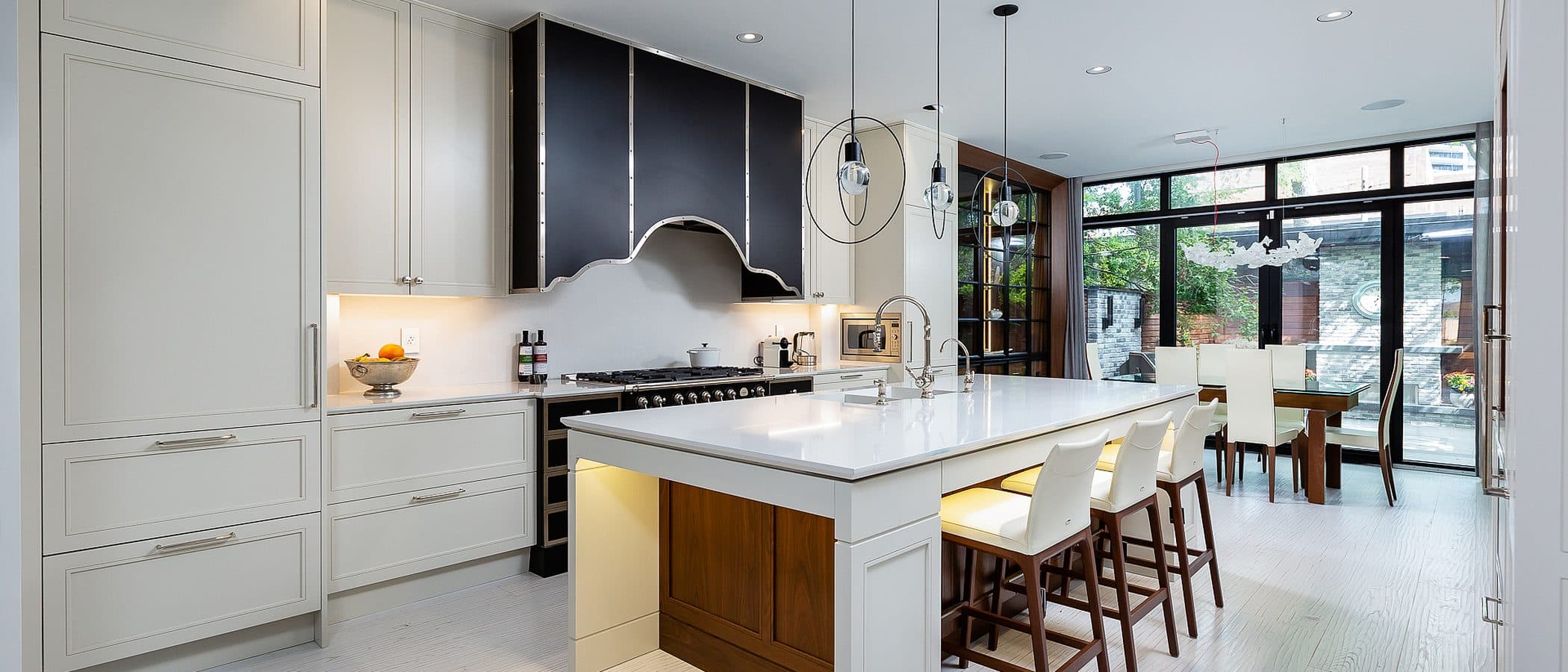 Open concept kitchen with white cabinetry, black paneled range hood, waterfall island, and dining table beyond glass doors to the garden.