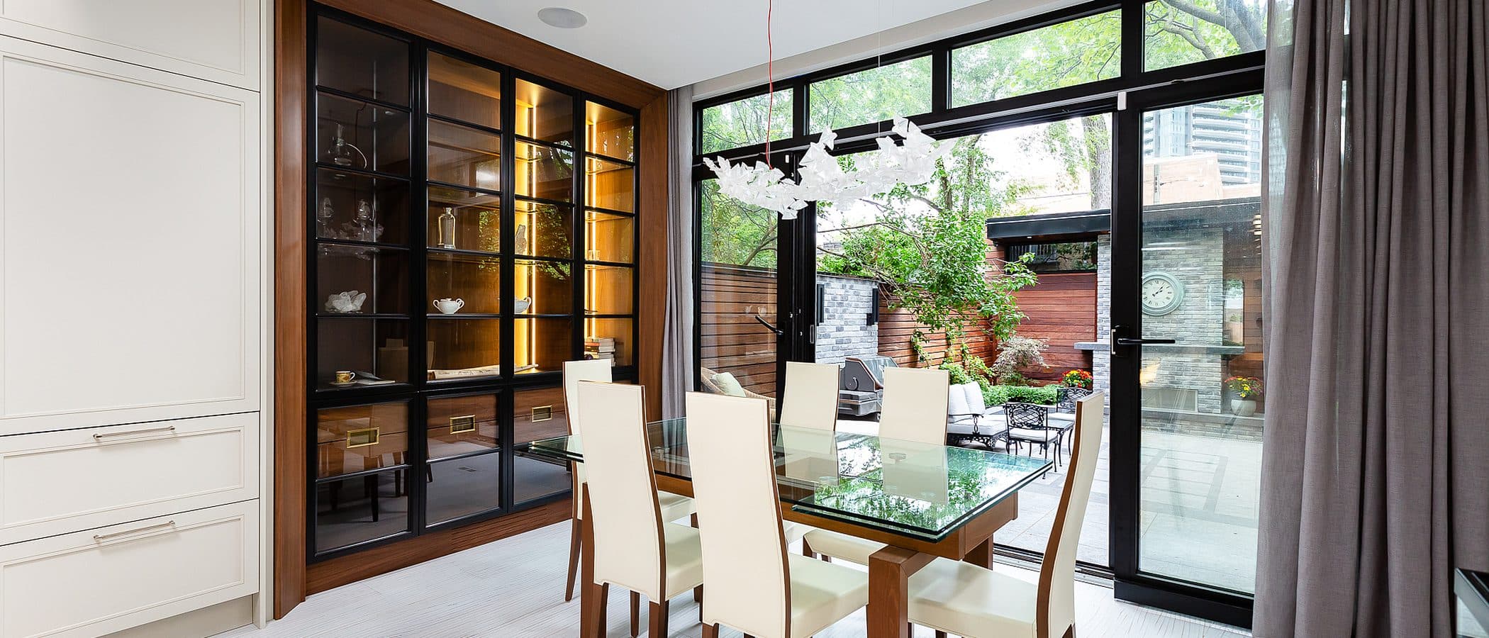 Dining room with glass table, upholstered chairs, and a walnut framed glass front cabinet lit from within, opening to a rear terrace.