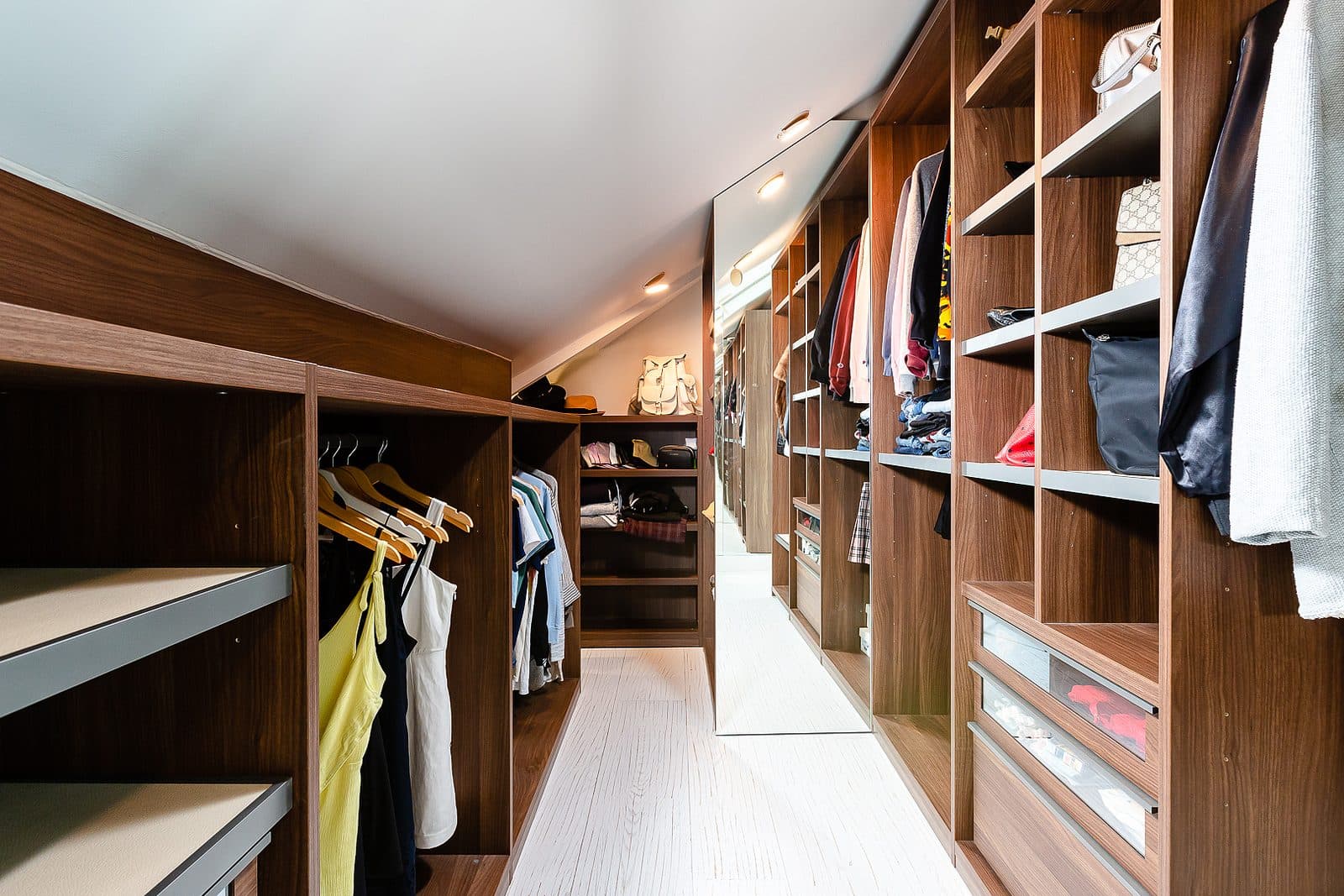 Walnut walk-in closet with open hanging bays, built-in drawers, and a mirrored end wall under a sloped ceiling.