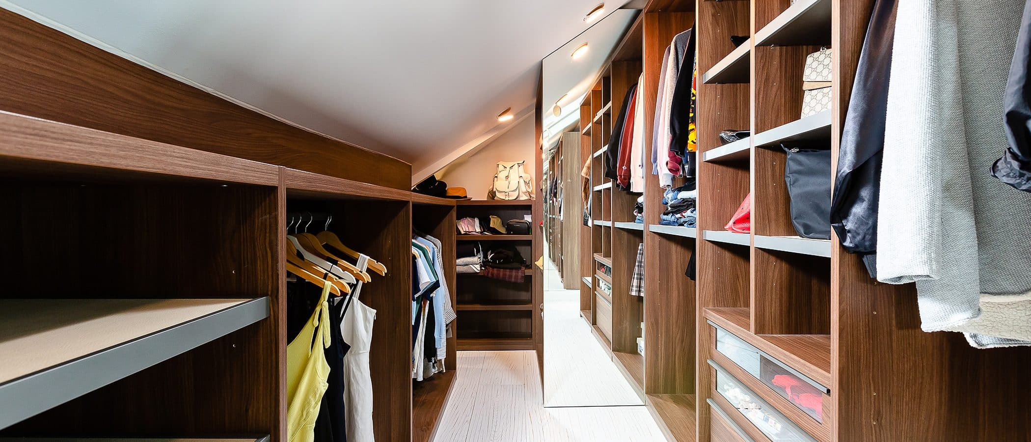 Walnut walk-in closet with open hanging bays, built-in drawers, and a mirrored end wall under a sloped ceiling.