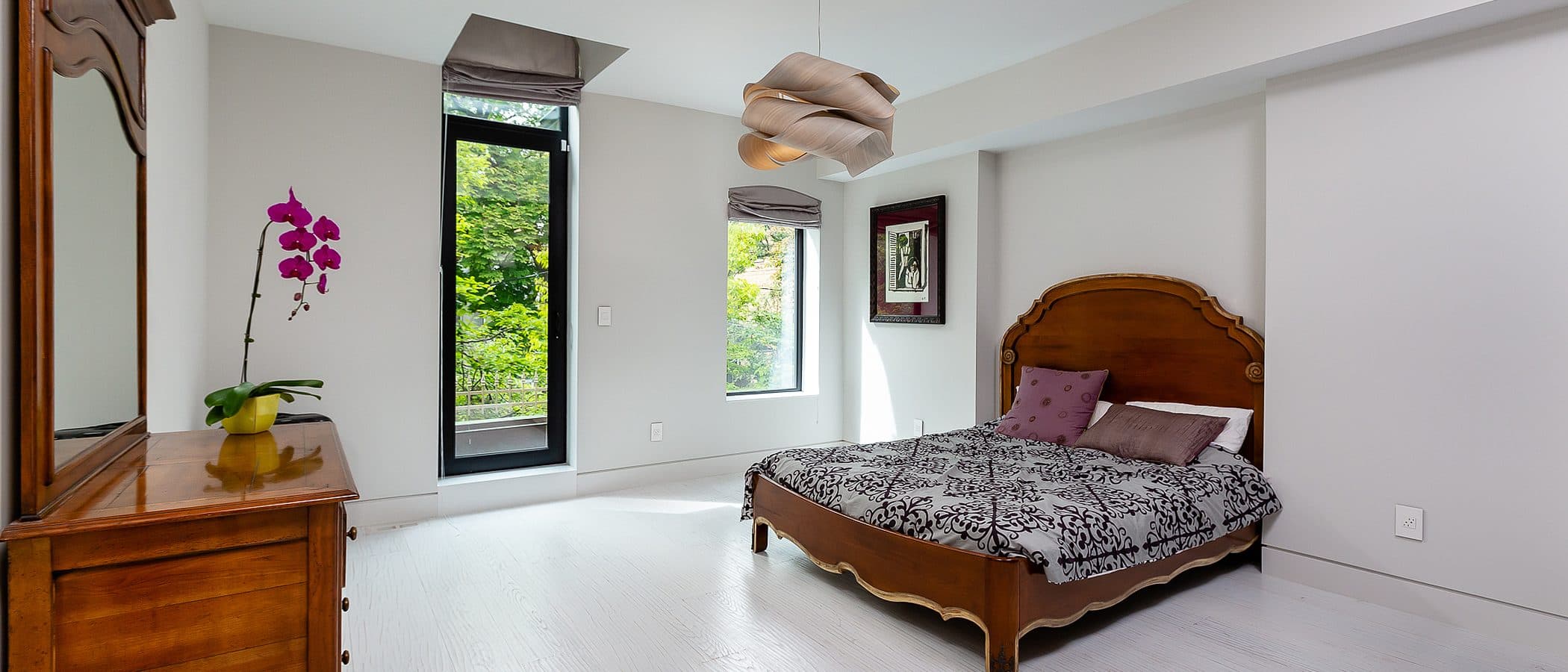 Bright bedroom with a carved wood antique bed frame, matching dresser, orchid, and a tall window overlooking trees.