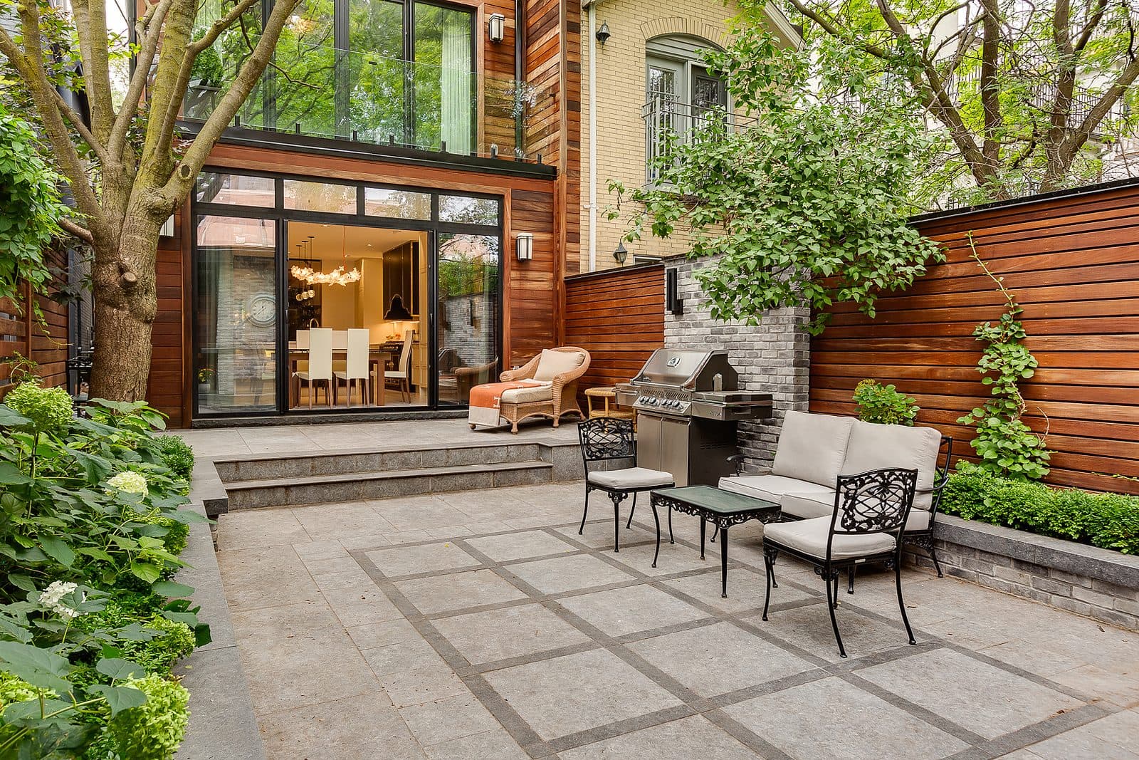 Rear glass and wood addition opening onto a stone patio with wrought iron furniture, an outdoor grill, and mature trees framing a brick neighbour.