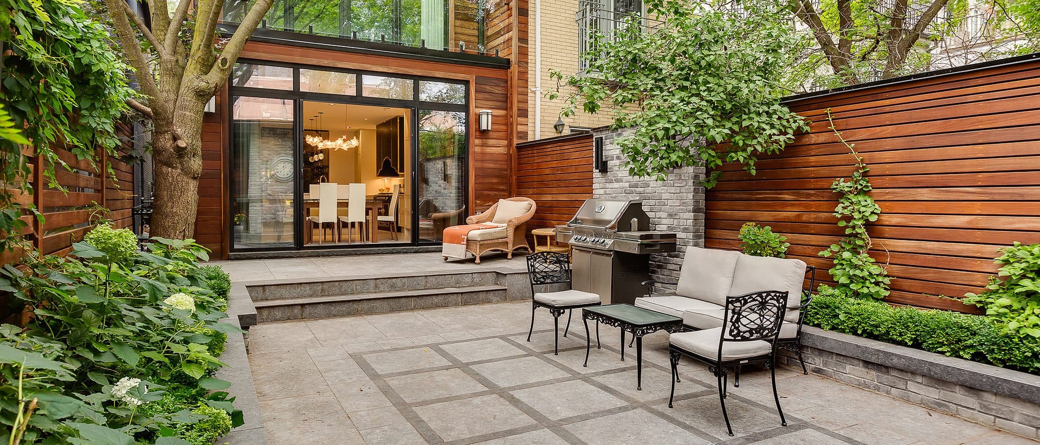 Rear glass and wood addition opening onto a stone patio with wrought iron furniture, an outdoor grill, and mature trees framing a brick neighbour.