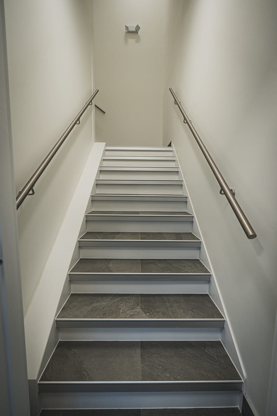 Straight on view down a stairwell with dual stainless steel handrails and dark stone tread stairs.